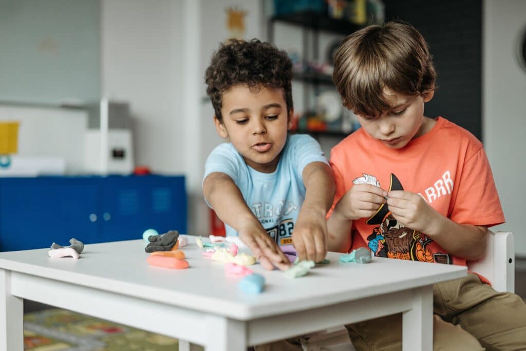 Play-doh is also an autism sensory toy. Pictured is two young boys sharing play-doh on a white table. autism toys