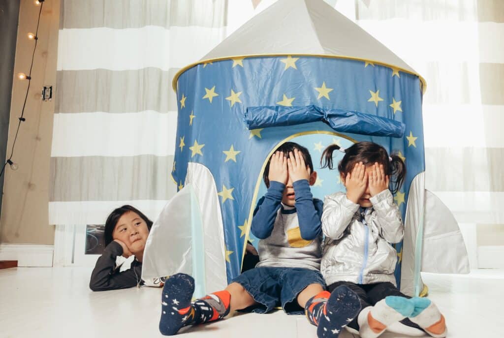 Three children playing hide and seek in an indoor tent. The first two children are covering their eyes and the third child is waiting for them to come find him. Hide and seek makes for a great game for autistic kids.