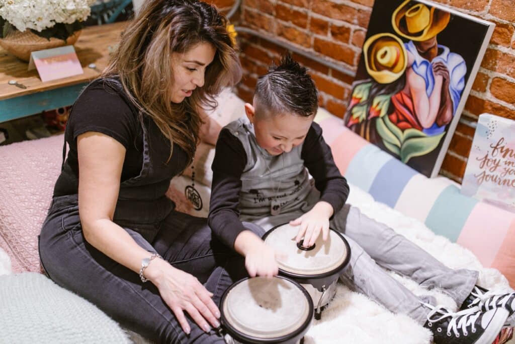 Young boy has fun drumming on bongos while his mom laughs close by.