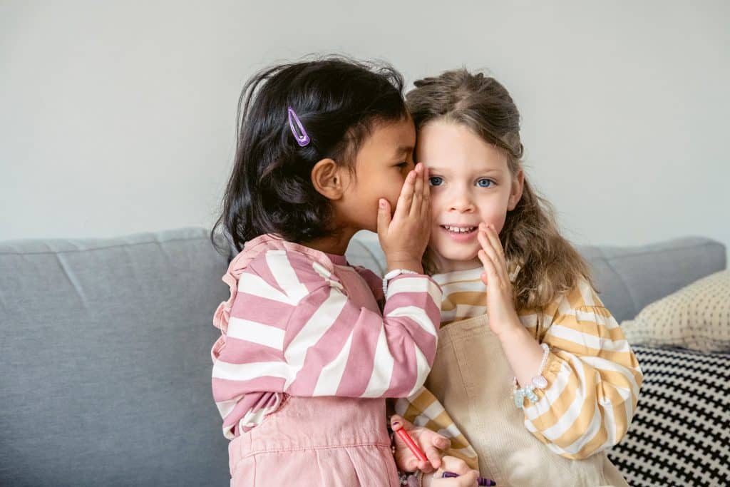 A little girl whispers in the ear of another little girl while playing telephone which is one of many communication activities for kids.