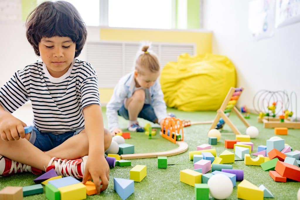 two kids play with various toys on the ground in the classroom.